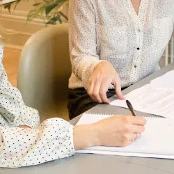 Doua femei lucreaza impreuna la completarea unor documente pe o masa bej