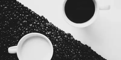 O cana alba cu cafea pe o masa alba si o cana alba cu lapte pe boabe negre de cafea