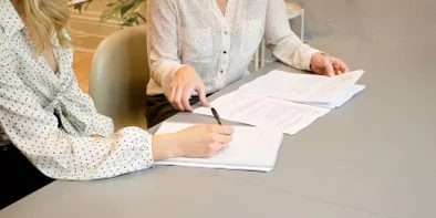 Doua femei lucreaza impreuna la completarea unor documente pe o masa bej