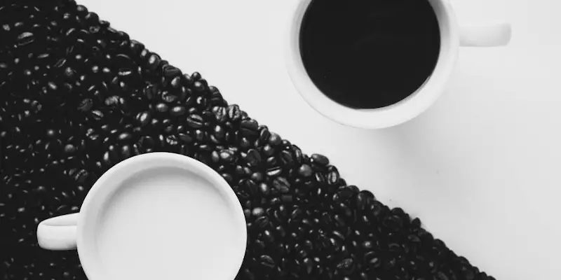 O cana alba cu cafea pe o masa alba si o cana alba cu lapte pe boabe negre de cafea