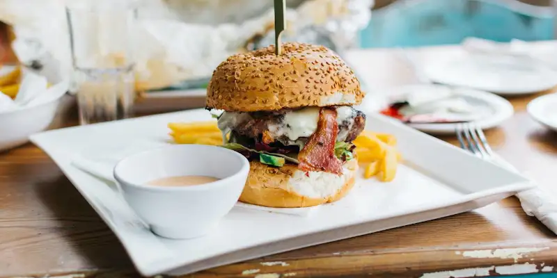 Un hamburger mare servit elegant pe o farfurie alba cu sos si cartofi prajiti